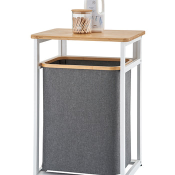 A modern laundry hamper features a white metal frame with a light wooden tabletop and a dark grey fabric bin suspended inside. Resting on top of the wooden surface are a clear glass jar filled with wooden clothespins and a white bottle of laundry detergent against a plain background.