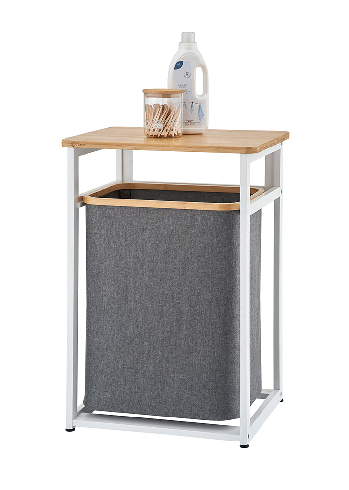 A modern laundry hamper features a white metal frame with a light wooden tabletop and a dark grey fabric bin suspended inside. Resting on top of the wooden surface are a clear glass jar filled with wooden clothespins and a white bottle of laundry detergent against a plain background.