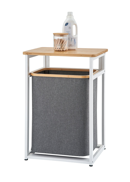 A modern laundry hamper features a white metal frame with a light wooden tabletop and a dark grey fabric bin suspended inside. Resting on top of the wooden surface are a clear glass jar filled with wooden clothespins and a white bottle of laundry detergent against a plain background.