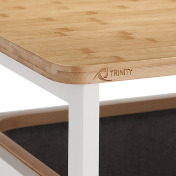 A detailed close-up shot showcases the rounded corner of a smooth, light-toned bamboo tabletop featuring a subtle "TRINITY" logo along the side. The wooden top rests on a clean white metal frame, with the rim of the dark gray fabric hamper visible underneath.