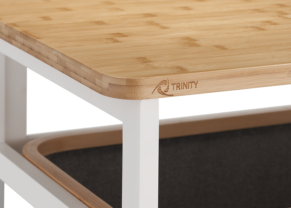 A detailed close-up shot showcases the rounded corner of a smooth, light-toned bamboo tabletop featuring a subtle "TRINITY" logo along the side. The wooden top rests on a clean white metal frame, with the rim of the dark gray fabric hamper visible underneath.