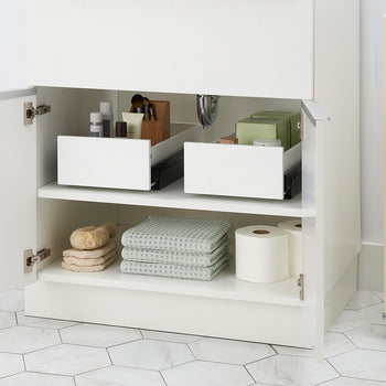 An open white bathroom cabinet features two pull-out drawer organizers filled with toiletries sitting partially closed on the top shelf. The bottom shelf is neatly organized with folded waffle-weave towels, bath sponges, and rolls of toilet paper.