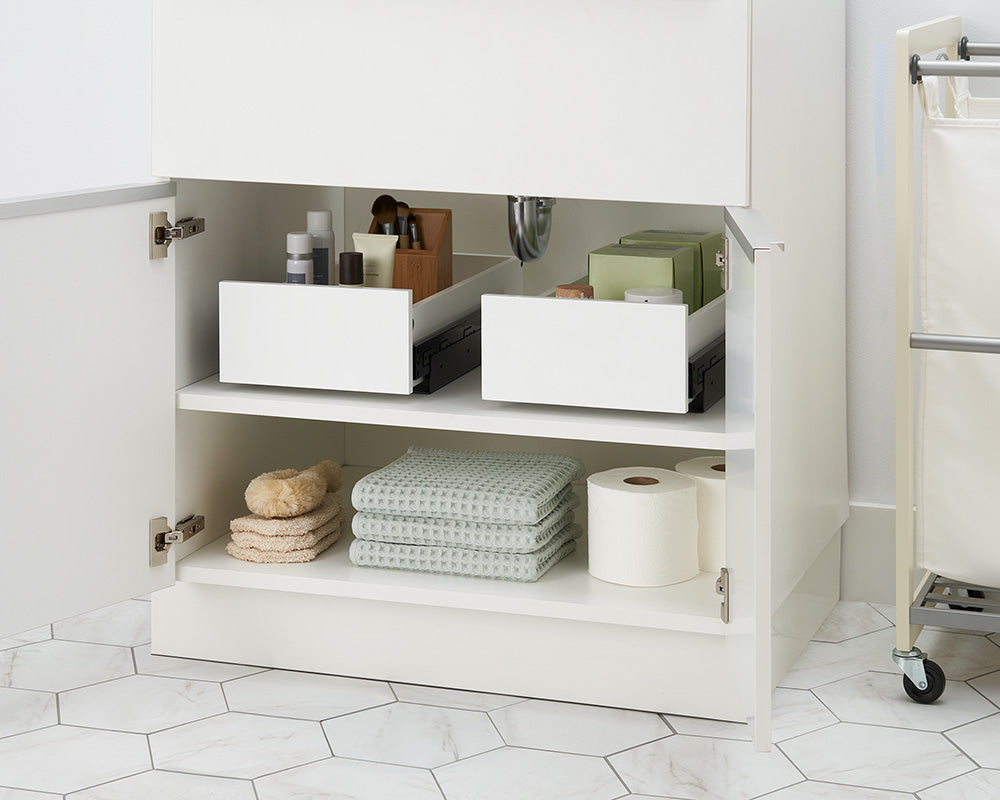 An open white bathroom cabinet features two pull-out drawer organizers filled with toiletries sitting partially closed on the top shelf. The bottom shelf is neatly organized with folded waffle-weave towels, bath sponges, and rolls of toilet paper.