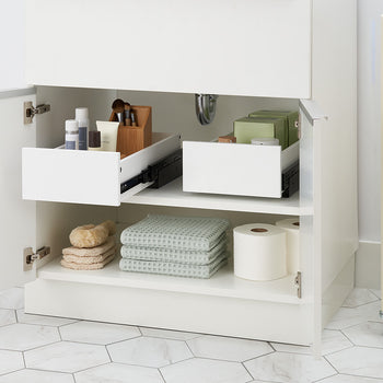 An open white bathroom vanity cabinet displays two pull-out drawer organizers mounted side by side on the top shelf. The left drawer holds cosmetic bottles and a wooden makeup brush caddy, while the lower shelf is neatly stocked with folded towels, bath sponges, and toilet paper rolls.
