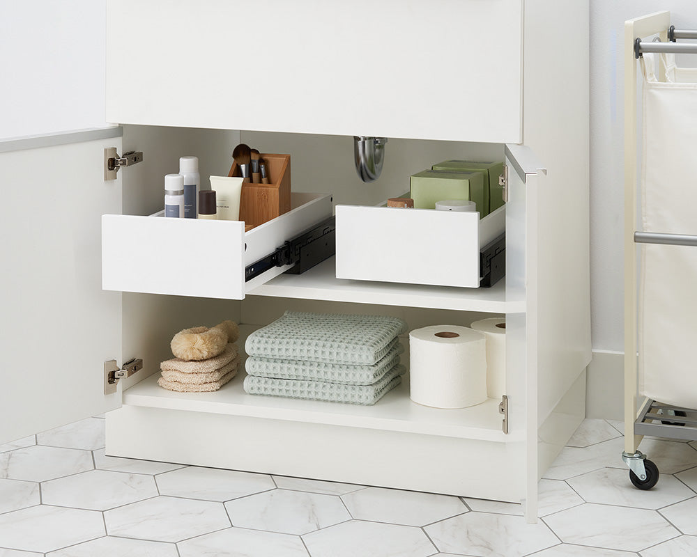 An open white bathroom vanity cabinet displays two pull-out drawer organizers mounted side by side on the top shelf. The left drawer holds cosmetic bottles and a wooden makeup brush caddy, while the lower shelf is neatly stocked with folded towels, bath sponges, and toilet paper rolls.
