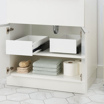 An open white bathroom cabinet reveals two empty pull-out organizer drawers installed side by side on the top shelf. Below them, folded waffle-weave towels, bath sponges, and toilet paper rolls are neatly arranged on the bottom shelf.