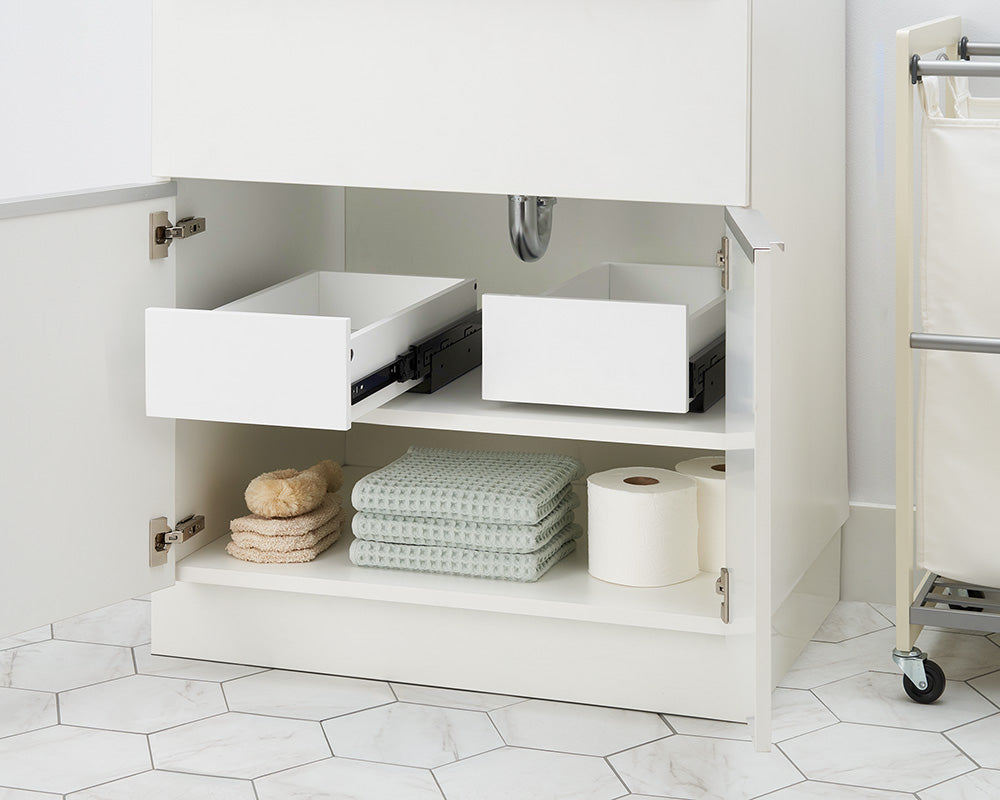 An open white bathroom cabinet reveals two empty pull-out organizer drawers installed side by side on the top shelf. Below them, folded waffle-weave towels, bath sponges, and toilet paper rolls are neatly arranged on the bottom shelf.
