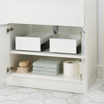 An open white bathroom cabinet reveals two empty pull-out organizer drawers resting partially open on the top shelf. The bottom shelf is organized with stacked waffle-weave towels, bath sponges, and toilet paper rolls.