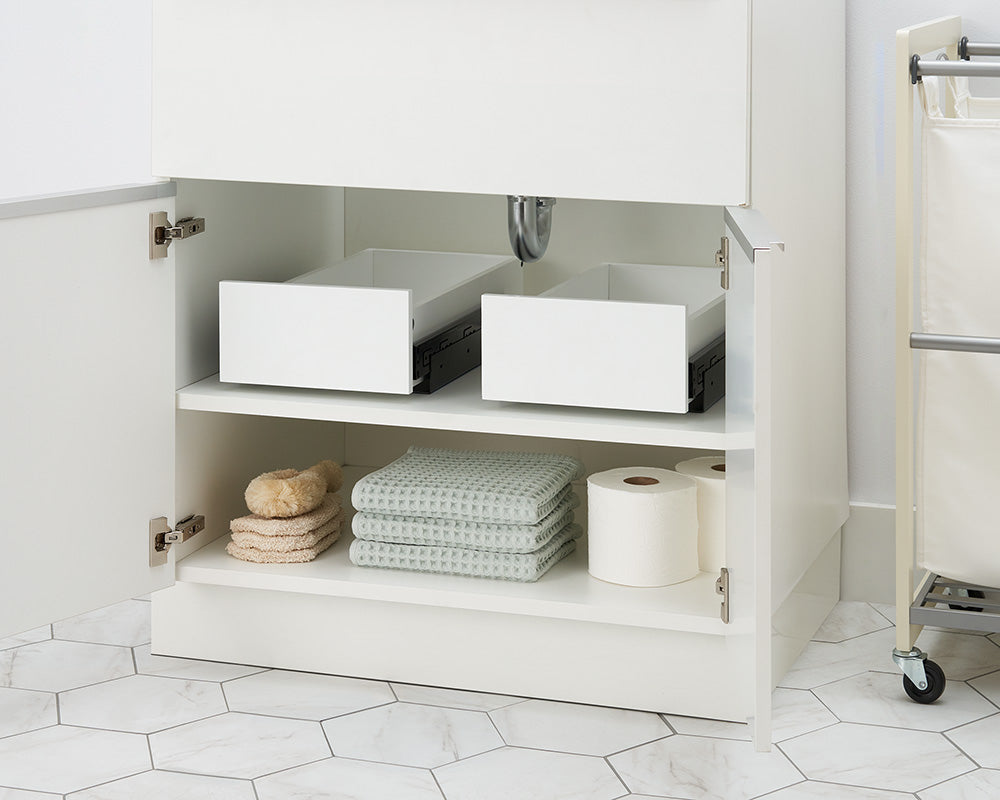 An open white bathroom cabinet reveals two empty pull-out organizer drawers resting partially open on the top shelf. The bottom shelf is organized with stacked waffle-weave towels, bath sponges, and toilet paper rolls.