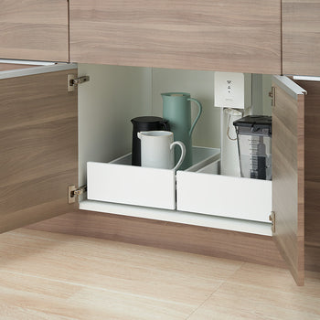 A low-angle view shows a wooden kitchen cabinet with both doors wide open, revealing two white pull-out drawer organizers inside. The left drawer holds a tall light-blue pitcher, a black kettle, and a white ceramic mug, while the right drawer contains a blender jar and a sparkling water maker.