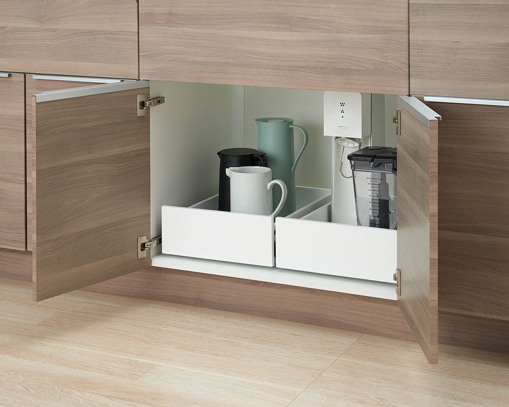 A low-angle view shows a wooden kitchen cabinet with both doors wide open, revealing two white pull-out drawer organizers inside. The left drawer holds a tall light-blue pitcher, a black kettle, and a white ceramic mug, while the right drawer contains a blender jar and a sparkling water maker.