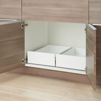 An open wooden kitchen cabinet is shown with two empty white pull-out drawers installed side-by-side inside. The interior of the cabinet is white, set above light wood-grain flooring.