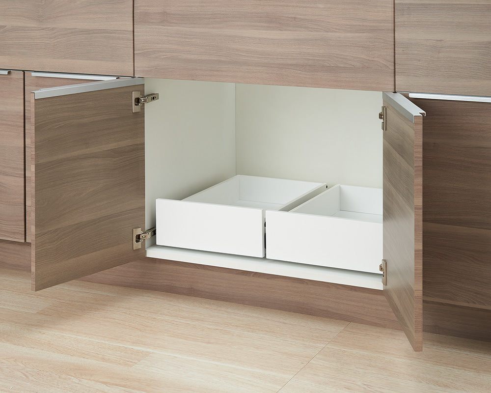 An open wooden kitchen cabinet is shown with two empty white pull-out drawers installed side-by-side inside. The interior of the cabinet is white, set above light wood-grain flooring.