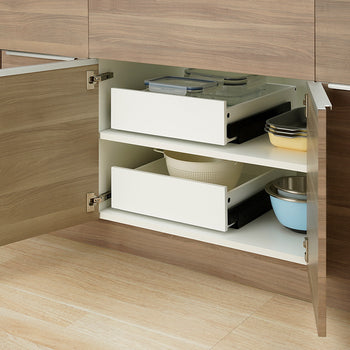 Two white pull-out organizer drawers are shown pushed fully inside an open wooden kitchen cabinet. The top drawer is filled with clear glass food containers, while the bottom drawer holds a colander, flanked by stacked baking dishes and mixing bowls on the shelves.