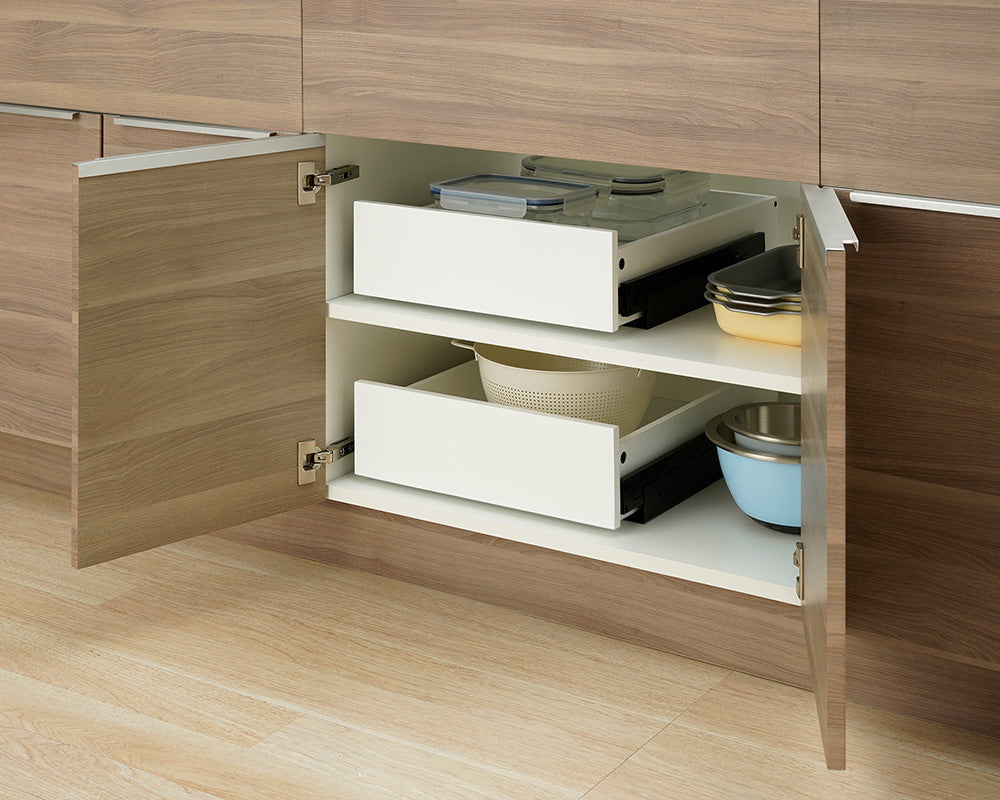 Two white pull-out organizer drawers are shown pushed fully inside an open wooden kitchen cabinet. The top drawer is filled with clear glass food containers, while the bottom drawer holds a colander, flanked by stacked baking dishes and mixing bowls on the shelves.