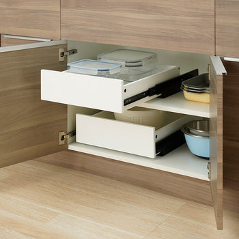 An open wooden kitchen cabinet displays two stacked white pull-out drawers mounted on different internal shelf levels. The top drawer is pulled outward to reveal clear glass food containers, while stacked baking dishes and mixing bowls sit alongside the drawers on the cabinet shelves.