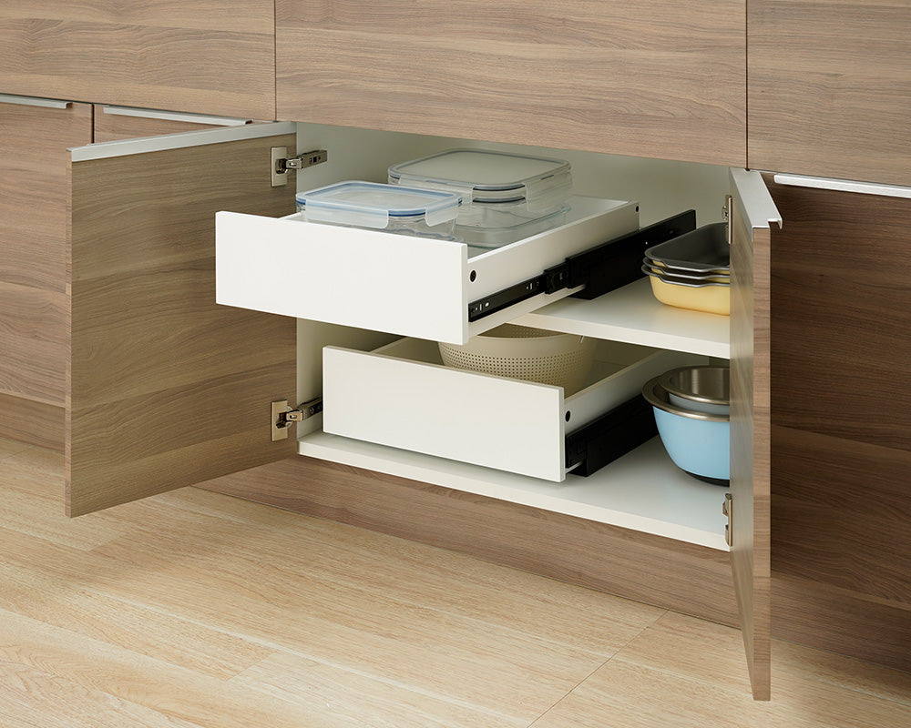 An open wooden kitchen cabinet displays two stacked white pull-out drawers mounted on different internal shelf levels. The top drawer is pulled outward to reveal clear glass food containers, while stacked baking dishes and mixing bowls sit alongside the drawers on the cabinet shelves.