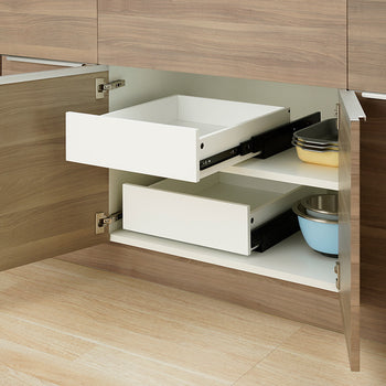 An open wooden kitchen cabinet reveals two empty white pull-out drawers installed on separate interior shelf levels. The top drawer is extended along black metal side rails, while nesting bowls and baking dishes sit directly on the shelves next to the drawers.