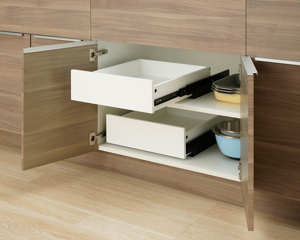 An open wooden kitchen cabinet reveals two empty white pull-out drawers installed on separate interior shelf levels. The top drawer is extended along black metal side rails, while nesting bowls and baking dishes sit directly on the shelves next to the drawers.