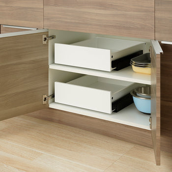 Two empty white pull-out drawers sit fully retracted inside an open wooden kitchen cabinet. Stacked baking pans and mixing bowls are neatly stored on the shelves beside the drawers.