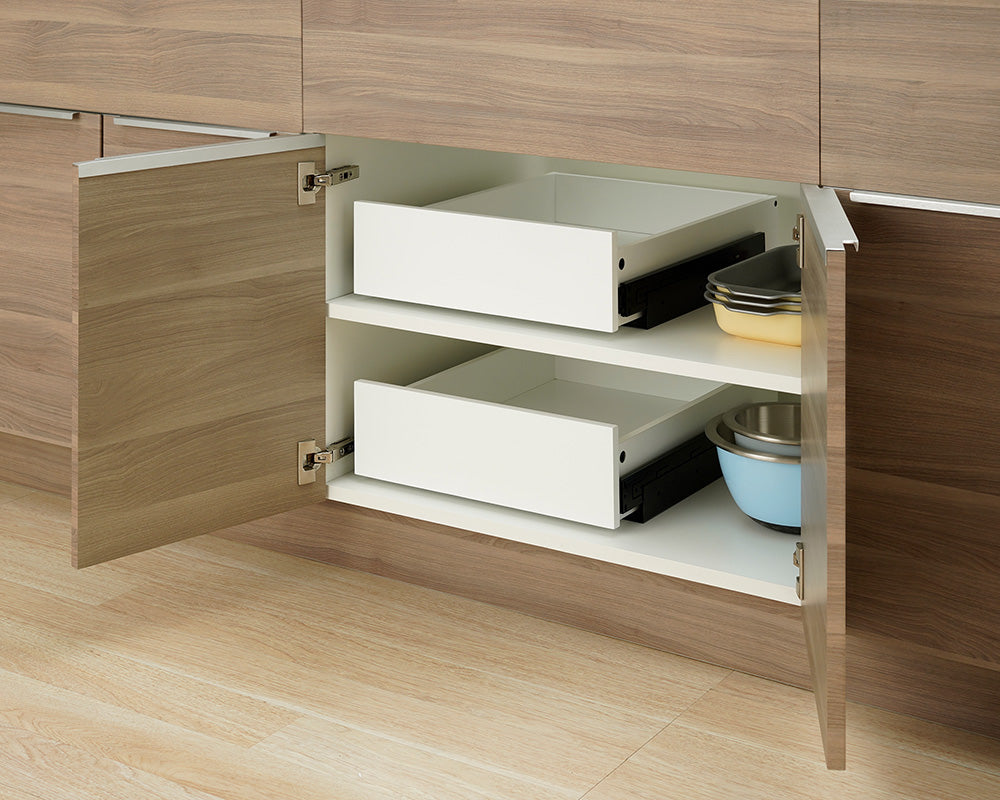 Two empty white pull-out drawers sit fully retracted inside an open wooden kitchen cabinet. Stacked baking pans and mixing bowls are neatly stored on the shelves beside the drawers.