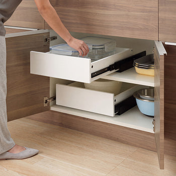 A person reaches into an open wooden kitchen cabinet with two stacked white pull-out drawers installed on different shelf levels. The extended top drawer holds glass food storage containers, while the lower drawer and surrounding shelves store mixing bowls, colanders, and baking pans.