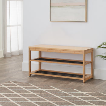 A light wood shoe storage bench with two black wire shelves sits against a white wall in a modern entryway. A patterned brown and white rug lies in front of the bench, and a potted plant sits next to it.