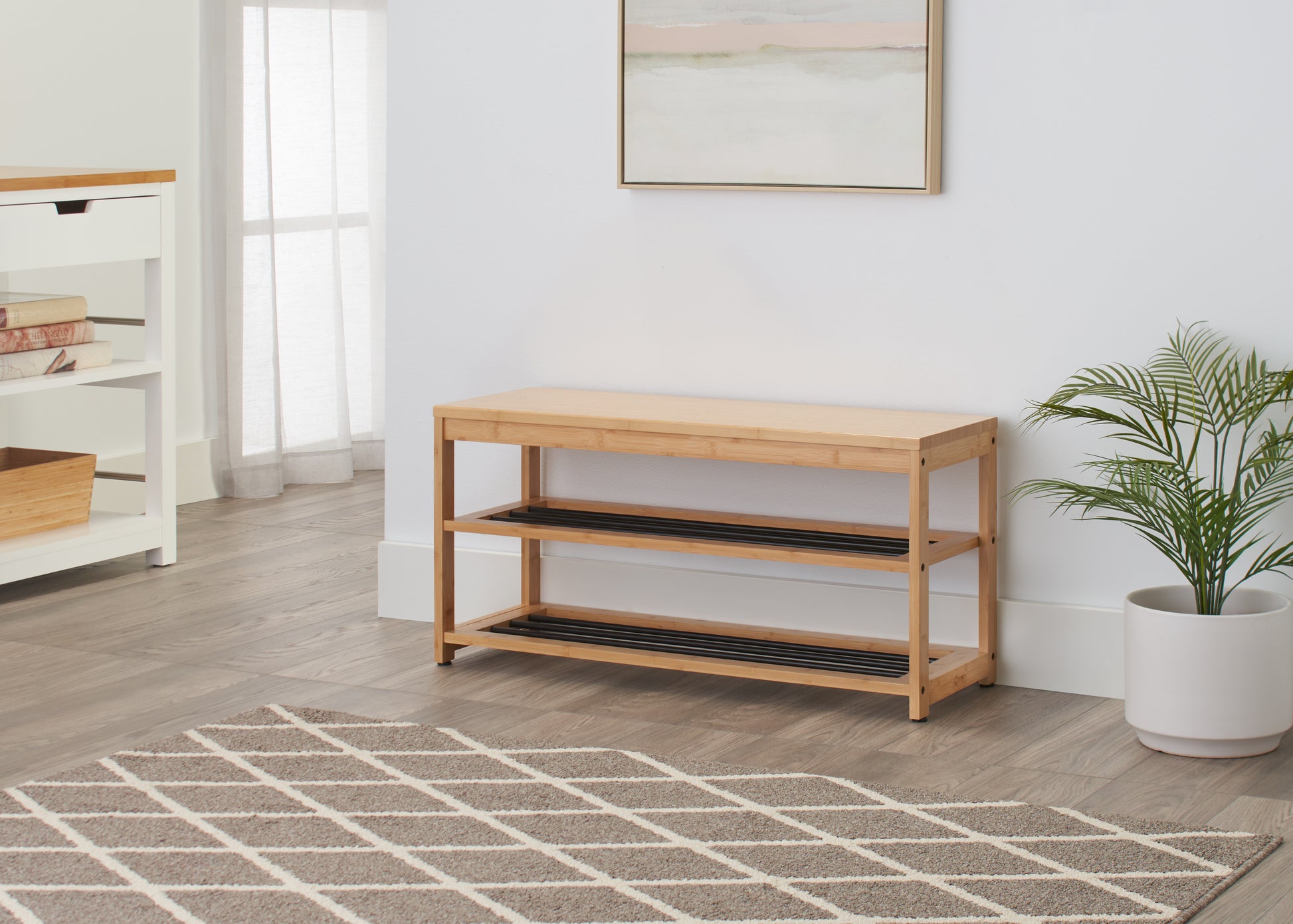 A light wood shoe storage bench with two black wire shelves sits against a white wall in a modern entryway. A patterned brown and white rug lies in front of the bench, and a potted plant sits next to it.