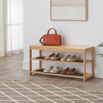 A light wood bench with two shelves sits on a geometric patterned rug in a bright room. The top of the bench holds a brown leather purse, and the shelves below are filled with several pairs of casual shoes.