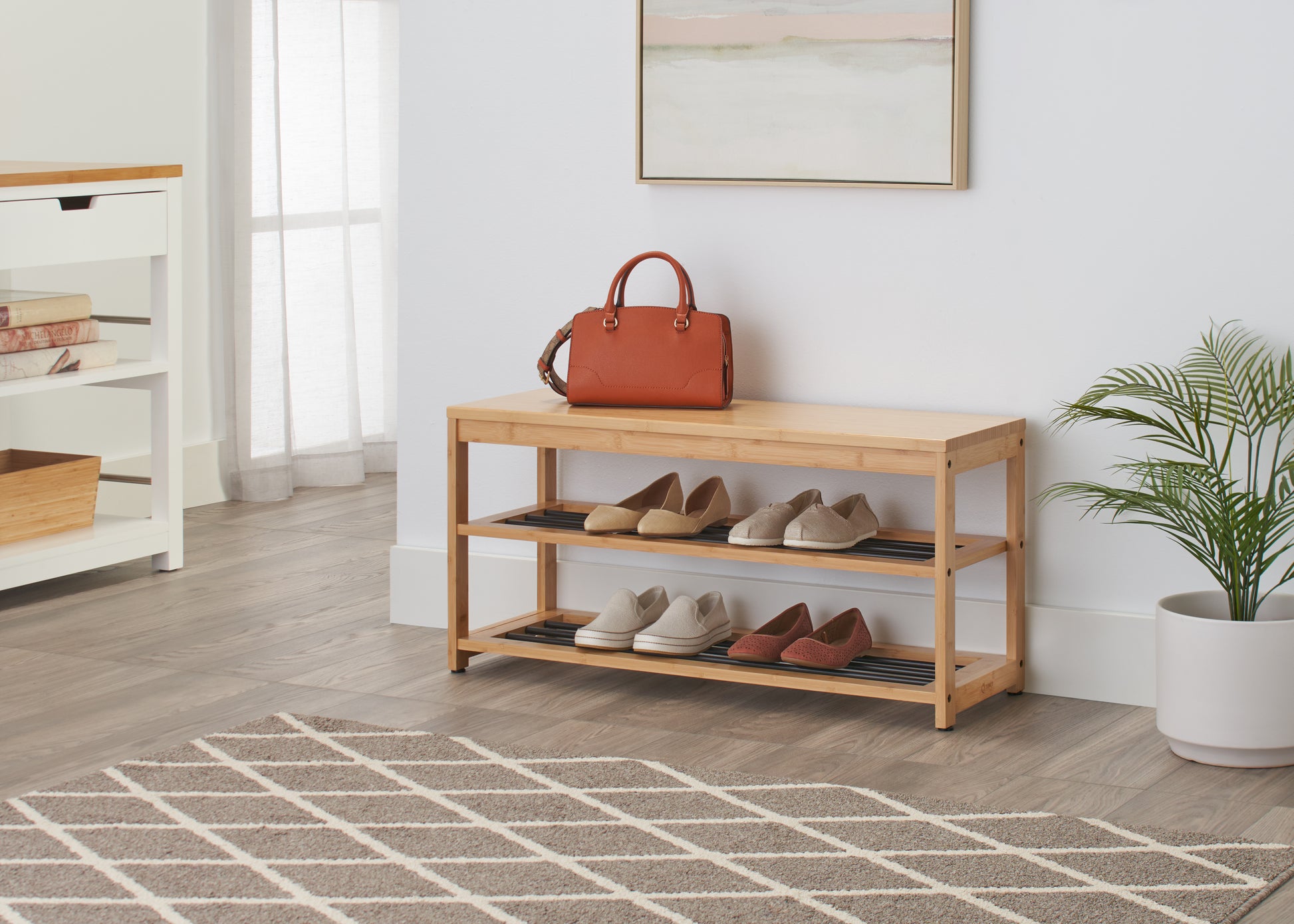 A light wood bench with two shelves sits on a geometric patterned rug in a bright room. The top of the bench holds a brown leather purse, and the shelves below are filled with several pairs of casual shoes.