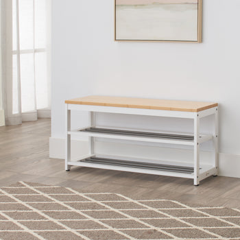 A wooden bench with a white metal frame sits in a hallway. It has two lower shelves with metal slats for shoe storage and is placed on a brown and white geometric patterned rug.