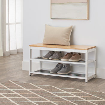 A shoe storage bench is placed in a light-filled entryway. The bench has a light wood top and two white metal racks below holding several pairs of shoes.