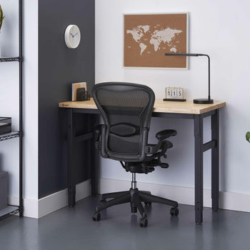 A modern home office setup features a light wood-topped desk with black metal legs and a black ergonomic office chair. The desk has a small lamp, a wooden block calendar showing 