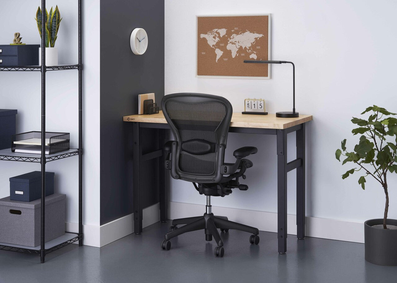A modern home office setup features a light wood-topped desk with black metal legs and a black ergonomic office chair. The desk has a small lamp, a wooden block calendar showing "15", and a few other small items, while a world map bulletin board hangs on the white wall behind it.