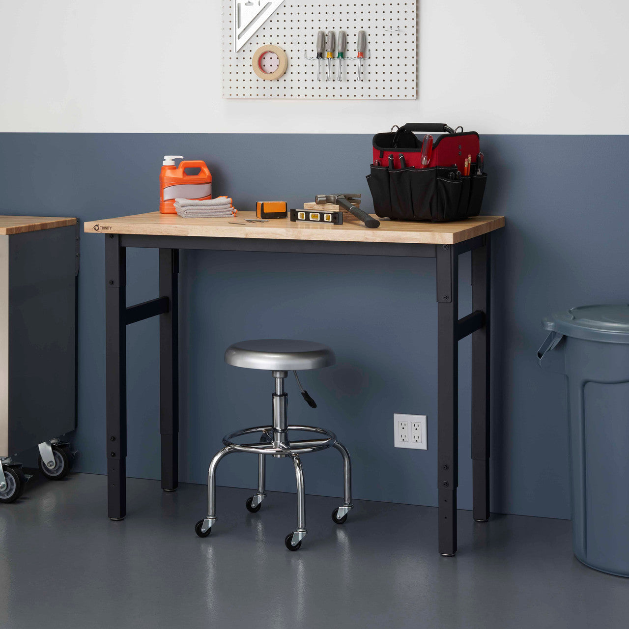 A light brown wooden-top work table with black metal legs stands against a two-toned wall, the lower half painted a dark blue. A silver rolling stool is tucked underneath the table, which holds various tools and supplies, including a red and black tool bag, orange cleaner, and a stack of cloths.