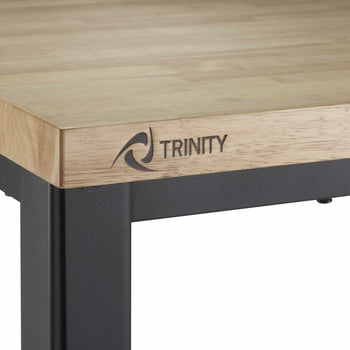 A close-up shot shows the corner of a Trinity brand work table. The table features a light-colored wooden top with visible grain and a black metal frame, including a leg and part of the side support. The 
