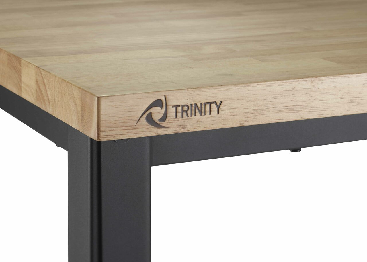A close-up shot shows the corner of a Trinity brand work table. The table features a light-colored wooden top with visible grain and a black metal frame, including a leg and part of the side support. The "TRINITY" logo is subtly printed on the edge of the wooden top.