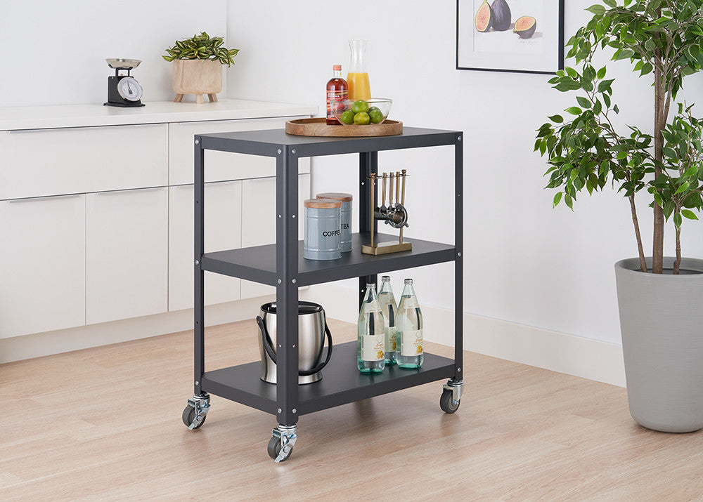 A gray metal utility cart with three shelves on wheels is positioned in a kitchen setting. The cart holds various items, including bottles, a bucket, and a tray with food.