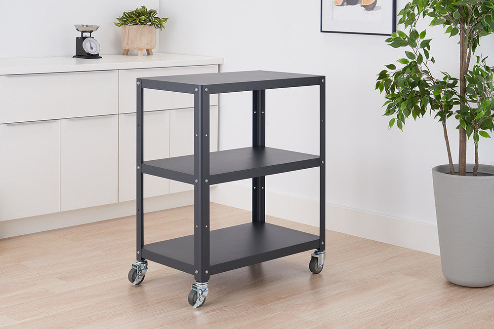 A dark gray metal utility cart with three shelves on wheels is positioned in a room with a white cabinet and a potted plant. The cart is empty, showcasing its simple design and functionality.