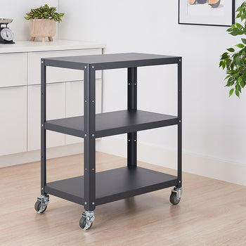 A dark gray metal utility cart with three shelves on wheels is positioned in a room with a white cabinet and a potted plant. The cart is empty, showcasing its simple design and functionality.