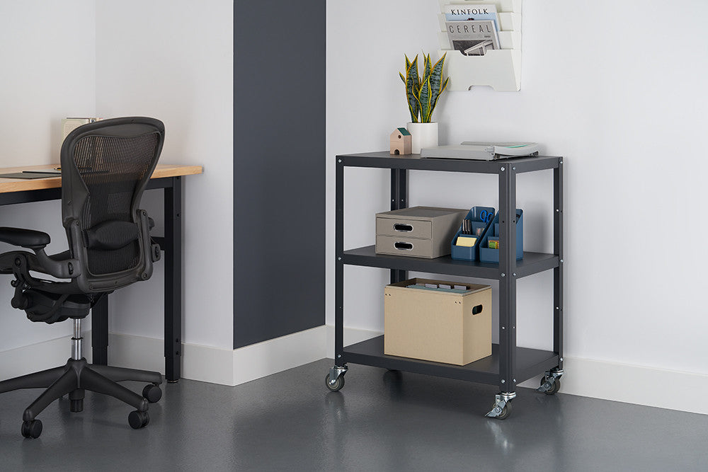 A dark gray metal utility cart with three shelves is positioned on a gray floor against a white wall with a gray accent stripe. The cart holds a laptop, storage boxes, and office supplies.