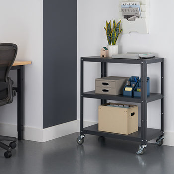 A dark gray metal utility cart with three shelves is positioned on a gray floor against a white wall with a gray accent stripe. The cart holds a laptop, storage boxes, and office supplies.
