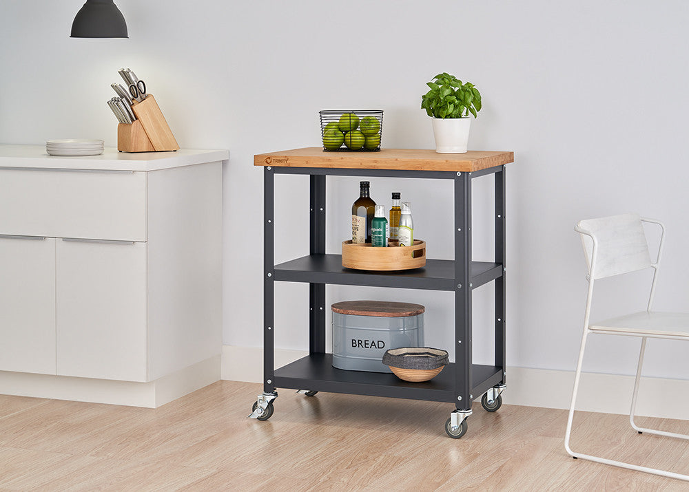 A kitchen cart with a light wood top and charcoal gray metal frame is centered in the image. It features two shelves and sits on caster wheels, with various kitchen items neatly arranged on each level.