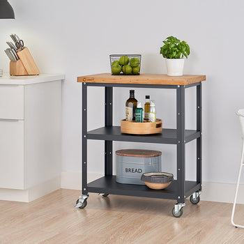 A kitchen cart with a light wood top and charcoal gray metal frame is centered in the image. It features two shelves and sits on caster wheels, with various kitchen items neatly arranged on each level.