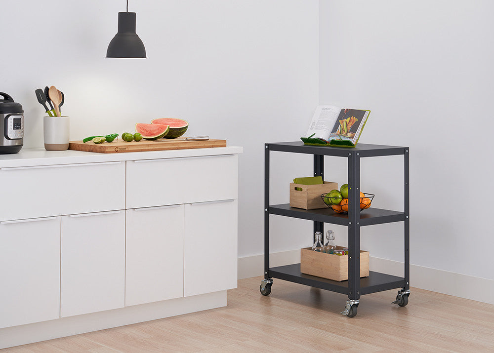 A dark gray metal utility cart with three shelves on wheels sits on a light-colored floor against a white wall in a kitchen setting. A cutting board with watermelon and limes rests on a white countertop to the left.