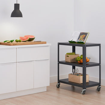 A dark gray metal utility cart with three shelves on wheels sits on a light-colored floor against a white wall in a kitchen setting. A cutting board with watermelon and limes rests on a white countertop to the left.