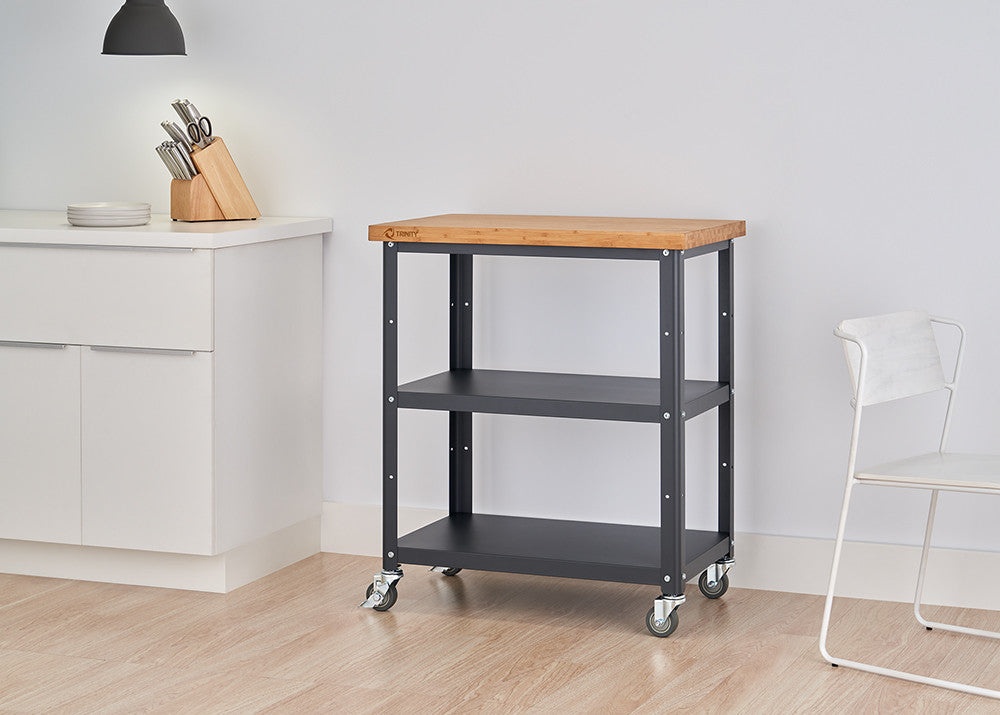 A medium shot of a kitchen cart with a light wood top and black metal frame with two shelves, positioned in a bright kitchen with white cabinets and a chair. The cart has wheels and is shown against a light-colored wood floor.