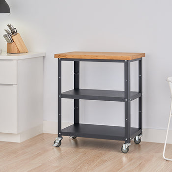 A medium shot of a kitchen cart with a light wood top and black metal frame with two shelves, positioned in a bright kitchen with white cabinets and a chair. The cart has wheels and is shown against a light-colored wood floor.