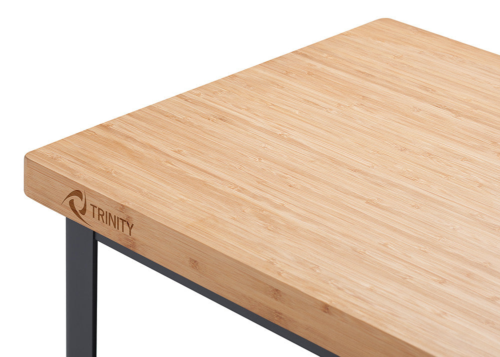 A close-up shot shows the corner of a light-colored bamboo countertop with the "TRINITY" logo subtly engraved on the edge. The countertop is attached to a dark metal frame, visible along the bottom and left edges of the image.