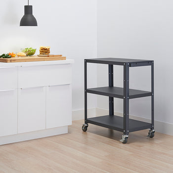 A kitchen scene shows white cabinets with minimalist handles and a dark grey, three-tiered rolling utility cart. On the counter, a wooden cutting board holds ingredients for tacos or similar food, illuminated by an overhead light.